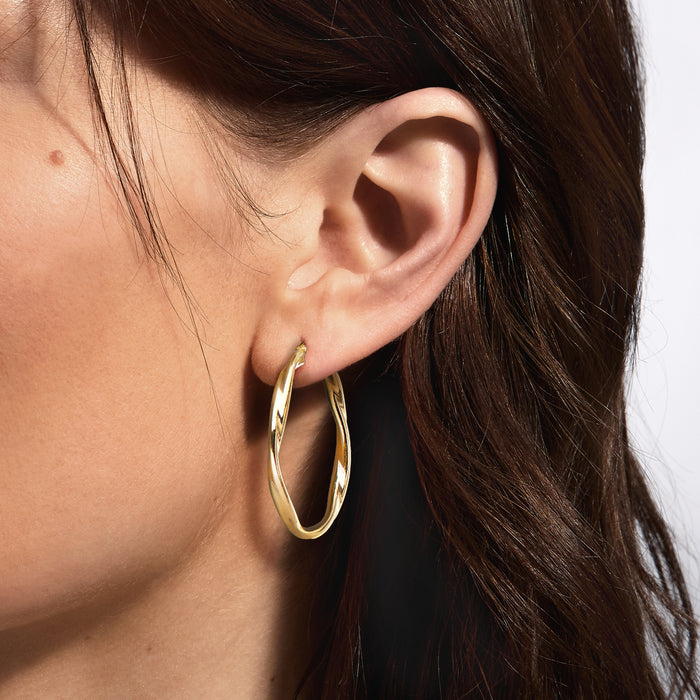 Twisted gold-tone hoop earrings worn on ear, oval hoop with shine