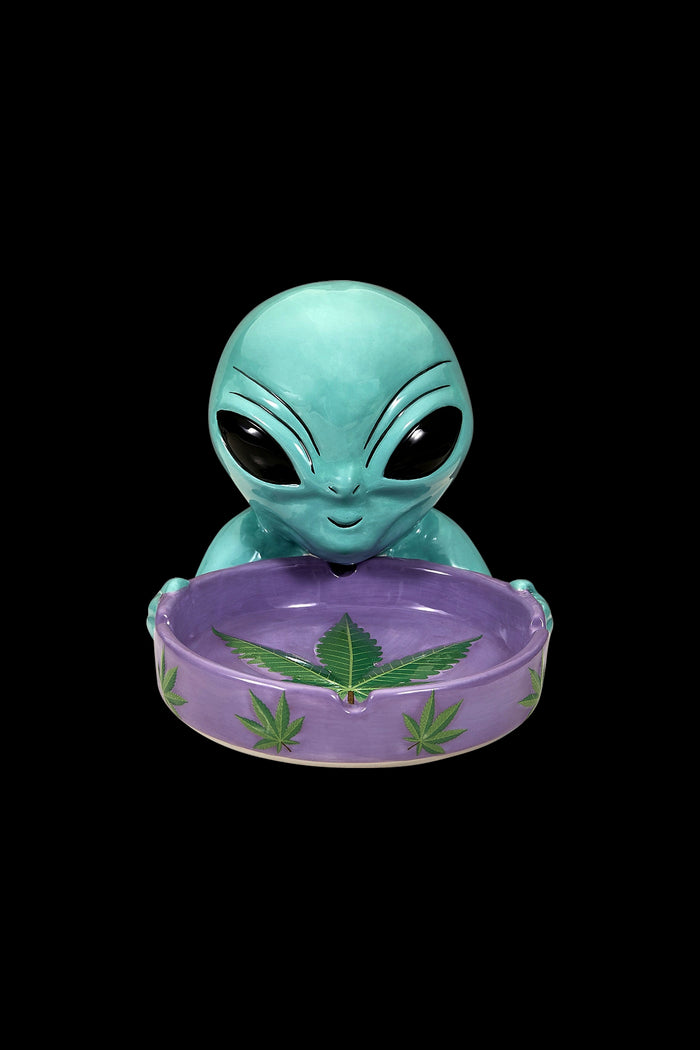 Turquoise alien head ashtray with cannabis leaf design on a purple base, isolated on black.