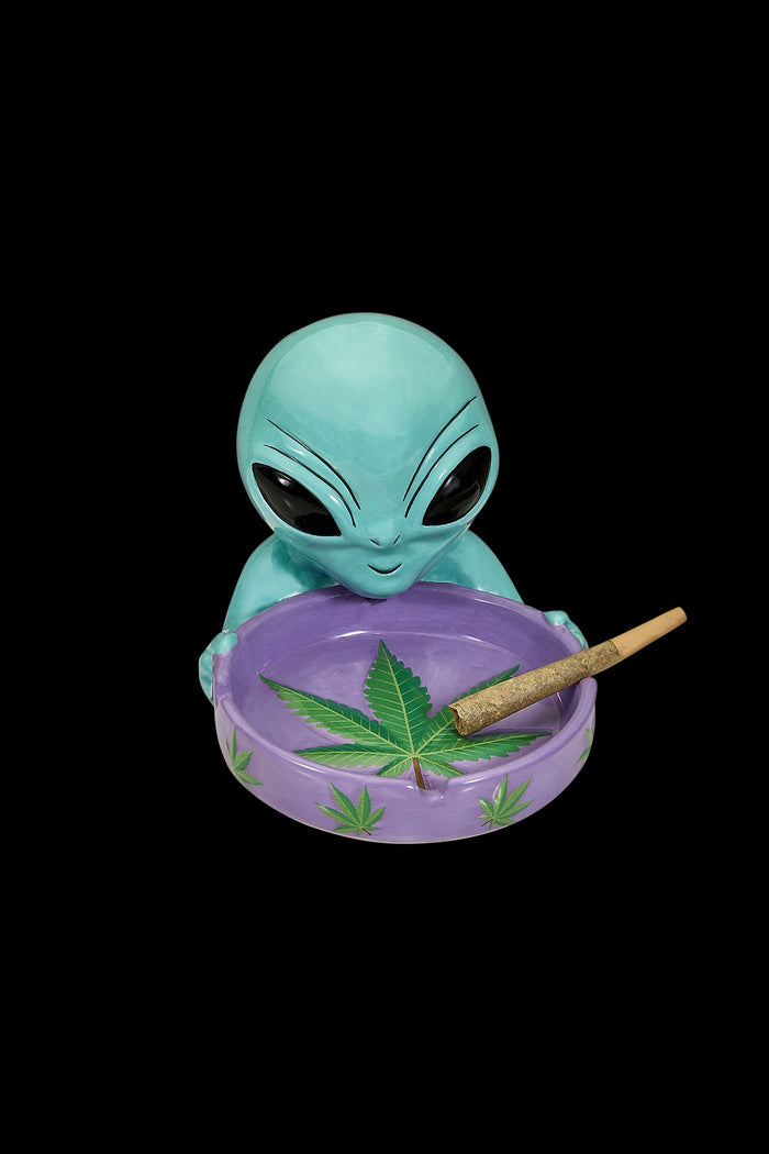 Ceramic ashtray shaped like an alien with a lit joint resting on a cannabis leaf design.